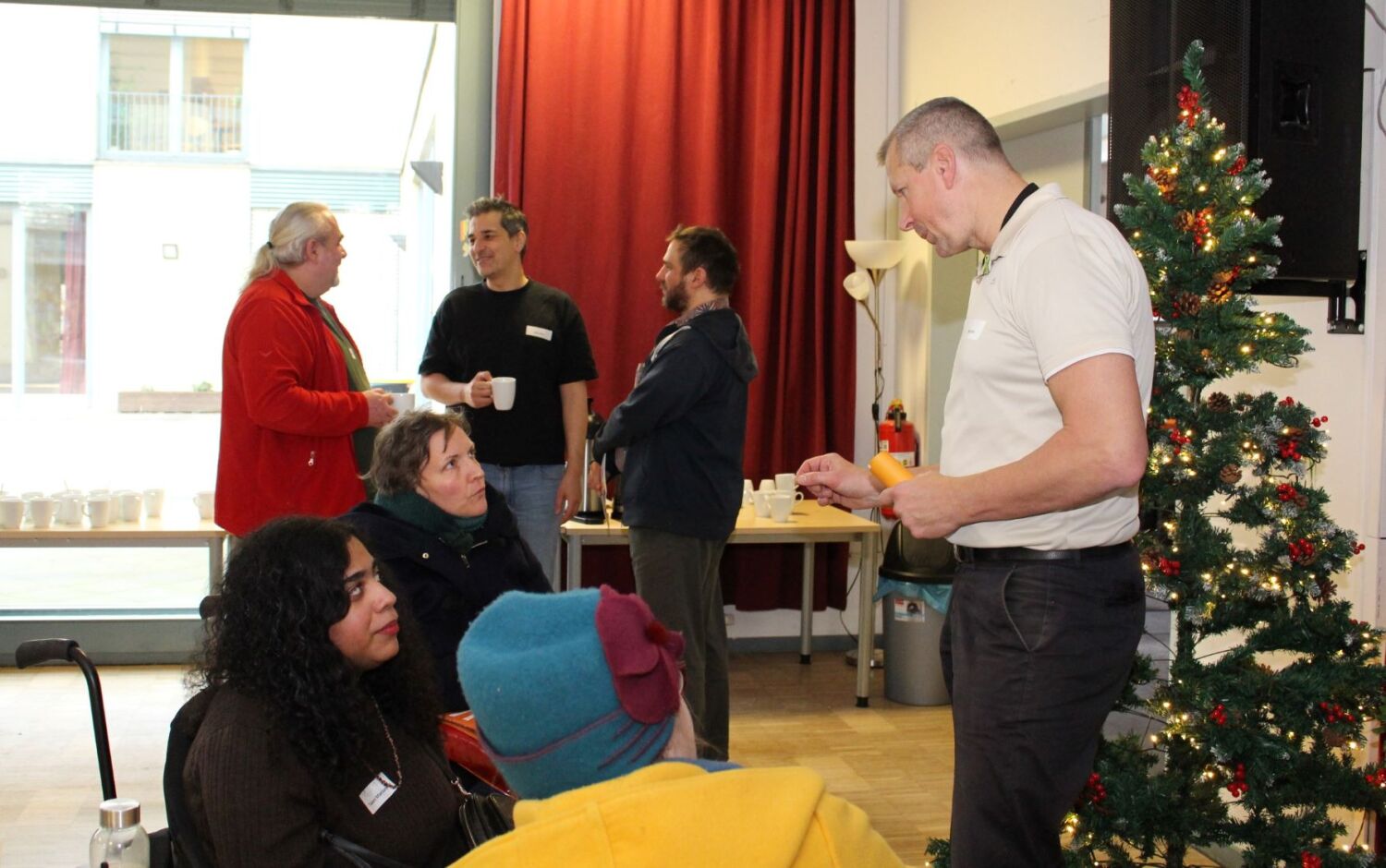 Ein Mann unterhält sich in einem Workshop mit drei sitzenden Frauen. Im Hintergrund steht eine Gruppe aus drei Männern im Gespräch.