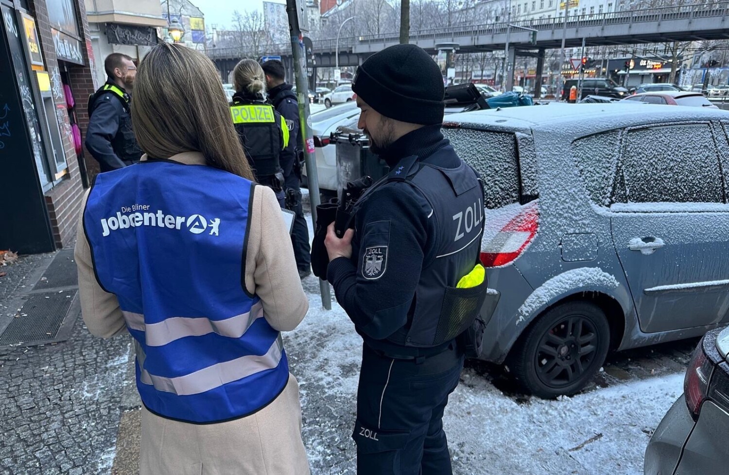 Eine Mitarbeiterin der Berliner Jobcenter und ein Beamter des Zolls von hinten vor einem Gebäude.