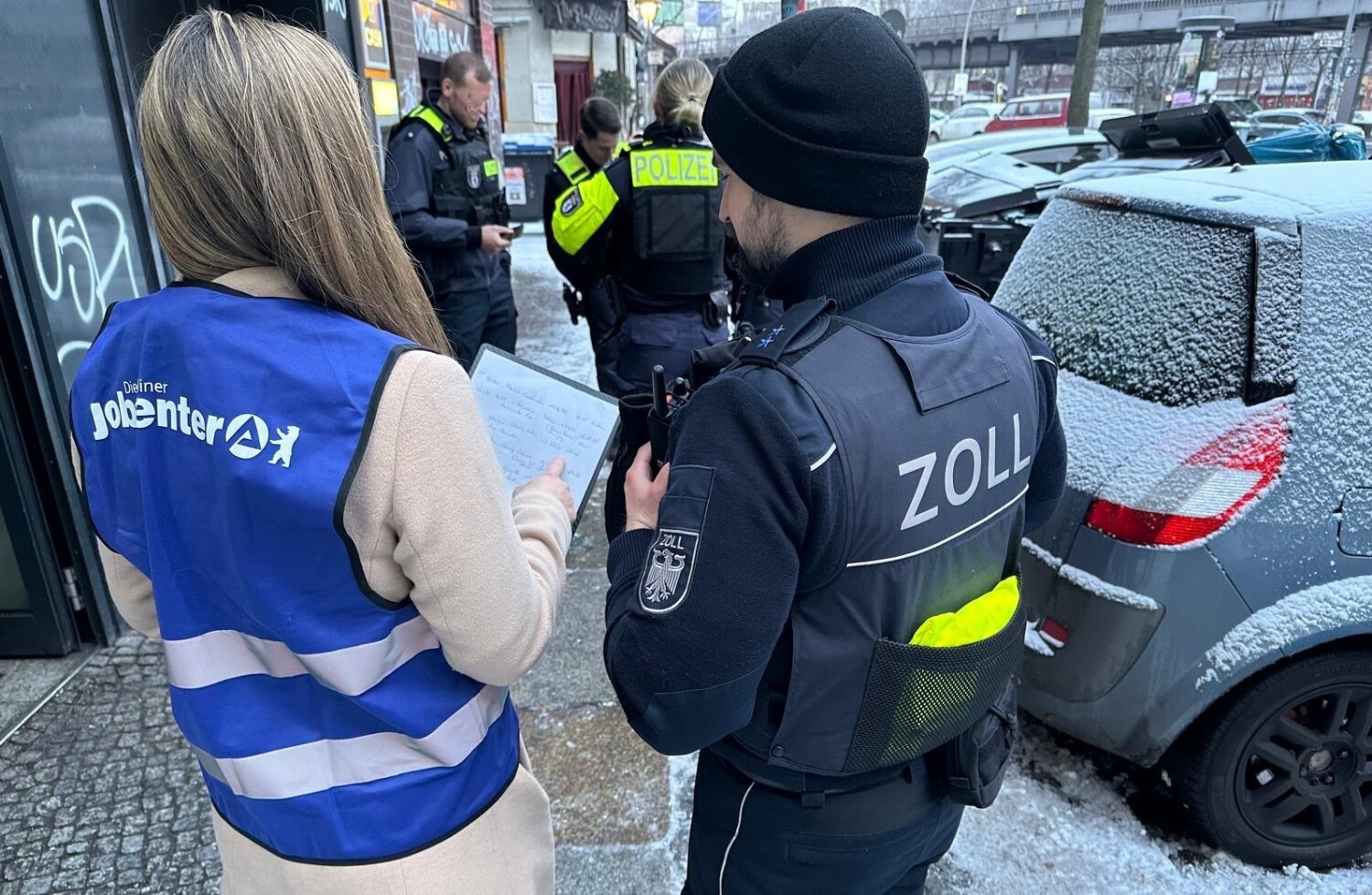 Eine Mitarbeiterin der Berliner Jobcenter und ein Beamter des Zolls von hinten vor einem Gebäude.