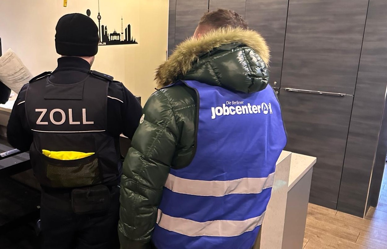 Beamte des Zolls und ein Mitarbeiter des Jobcenters sind von hinten in Räumlichkeit zu sehen.