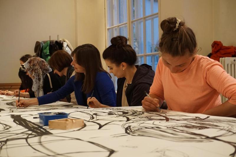 Nahaufnahme von 7 Frauen, die gemeinsam an der langen Tafel auf weiße Papierbahnen konzentriert zeichnen.