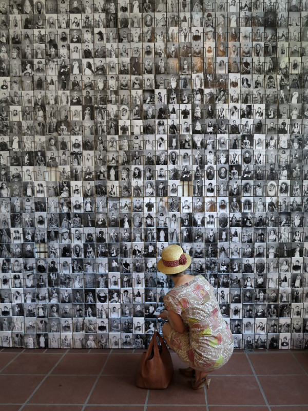 eine Frau hockt vor einer großen Wand, die vollständig mit schwarz-weiß Postkarten bedeckt ist, die Frauenporträts zeigen