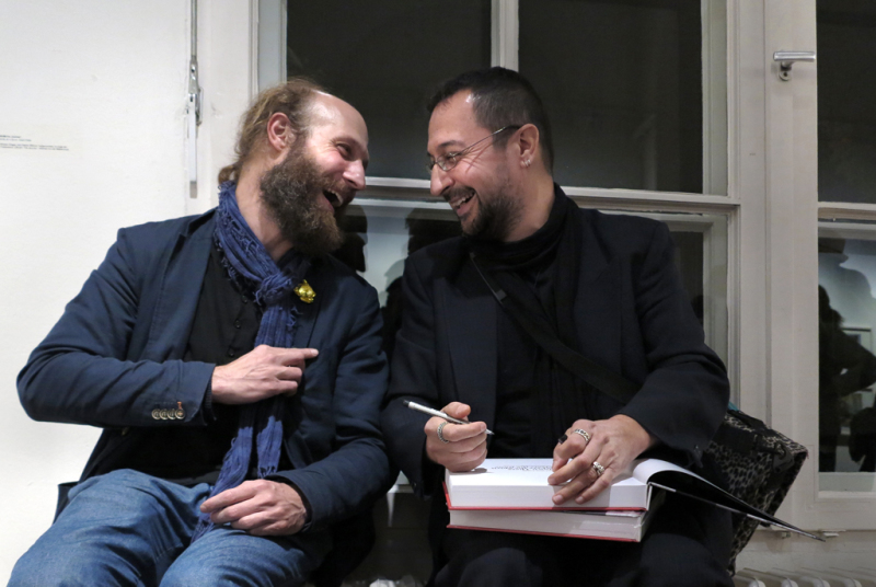 Nihad Nino Pusija mit seinem Galeristen im Gespräch, die zwei sitzen vor dem Fenster und lachen herzlich, während Pusija seine Publikation in den Händen hält und im Begriff ist, diese zu signieren.