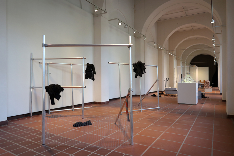  Im Galerieraum stehen Aluminiumstangen verteilt herum, die wie Gerüstteile aussehen. An manchen von ihnen hängen schwarze Kleidungsfetzen.