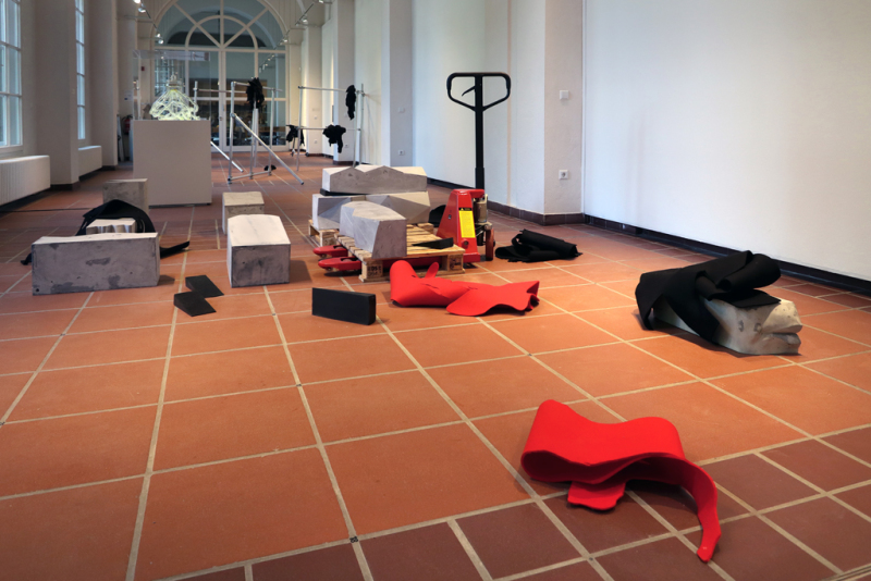 Ein Hubwagen mit einer Palette stehen mitten im Galerieraum. Darauf und in der Nähe liegen rote und schwarze Stoffteile und große graue Gipsblöcke, die mitunter als Einzelteile einer zusammenhängenden Skulptur wirken.