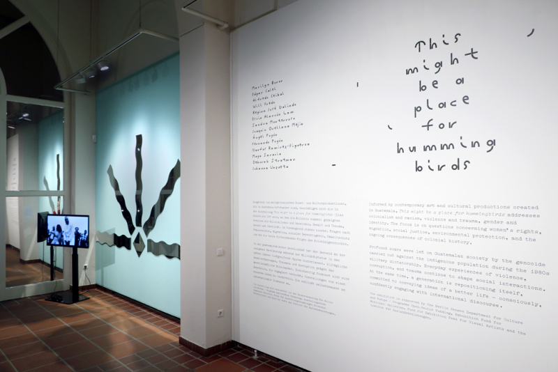Auf der rechten Seite ist der Wandtext zur Ausstellung "This might be a place for hummingbirds" zu sehen. Auf der linken Seite eine Installation bestehend aus 5 Sonnenförmig angeordneten Spiegeln auf einer türkis gestrichenen Wand von Maya Saravia.