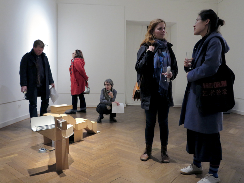 Drei Besucher betrachten aufmerksam die Arbeiten von Larrissa Fassler aus verschiedenen Perspektiven. Im Vordergrund sind zwei junge Frauen ind Gespräch vertieft. Die Gläser, die fast alle Personenn in den Händen halten, verweisen auf den Eröffnungsabend.