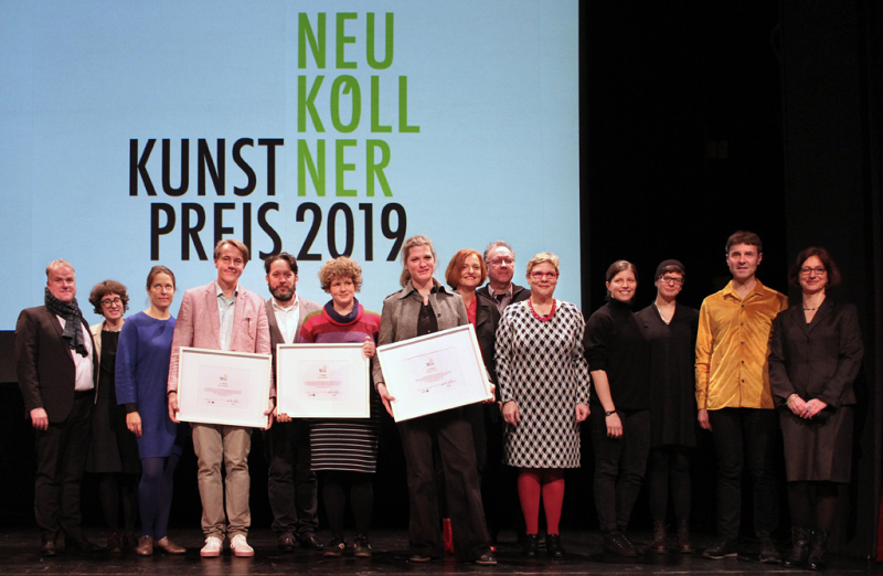Einer Reihe von Menschen auf einer Bühne vor der Projektion Neuköllner Kunstpreis 2019. Die drei Preisträger halten Rahmen mit Preis-Urkunden in den Händen.