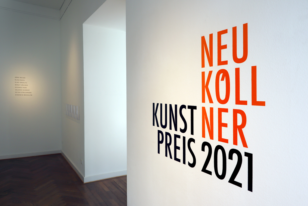 Der Titel der Ausstellung Neuköllner Kunstpreis steht in oranger Schrift auf der rechten Wand im Eingangsbereich der Galerie