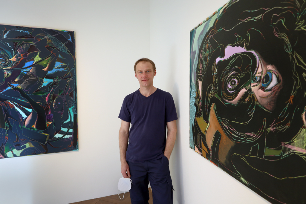 Arno Bojak steht zwischen zwei bunten Gemälden, die hinter ihm an der Wand hängen.