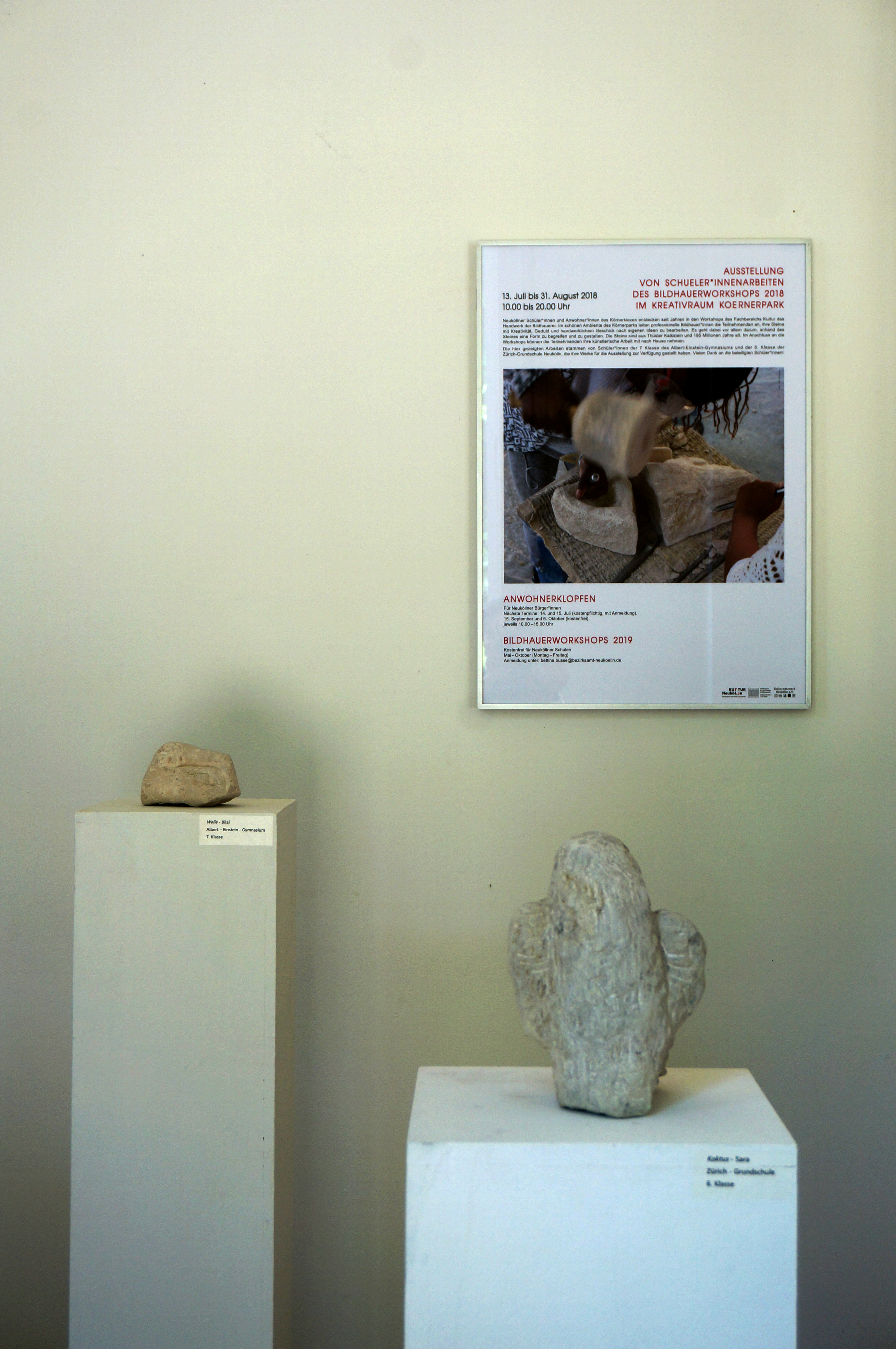 Im Vordergrund ein Kaktus aus Kalkstein auf einer weißen Stele, im Hintergrund eine weitere Steinskulptur auf einer Stele sowie ein Plakat an der Wand