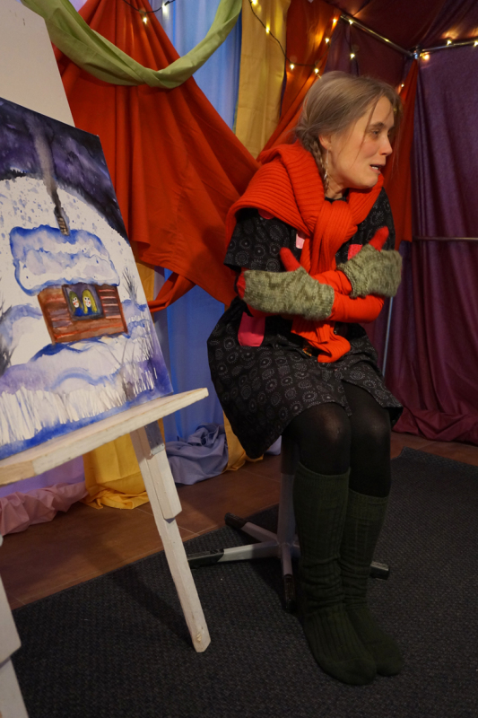 Eine Frau mit Handschuhen und Schal sitzt auf einem Hocker und hat die Arme frierend um sich geschlungen, neben ihr eine Staffelei mit einem Bild aus dem finnischen Märchen.