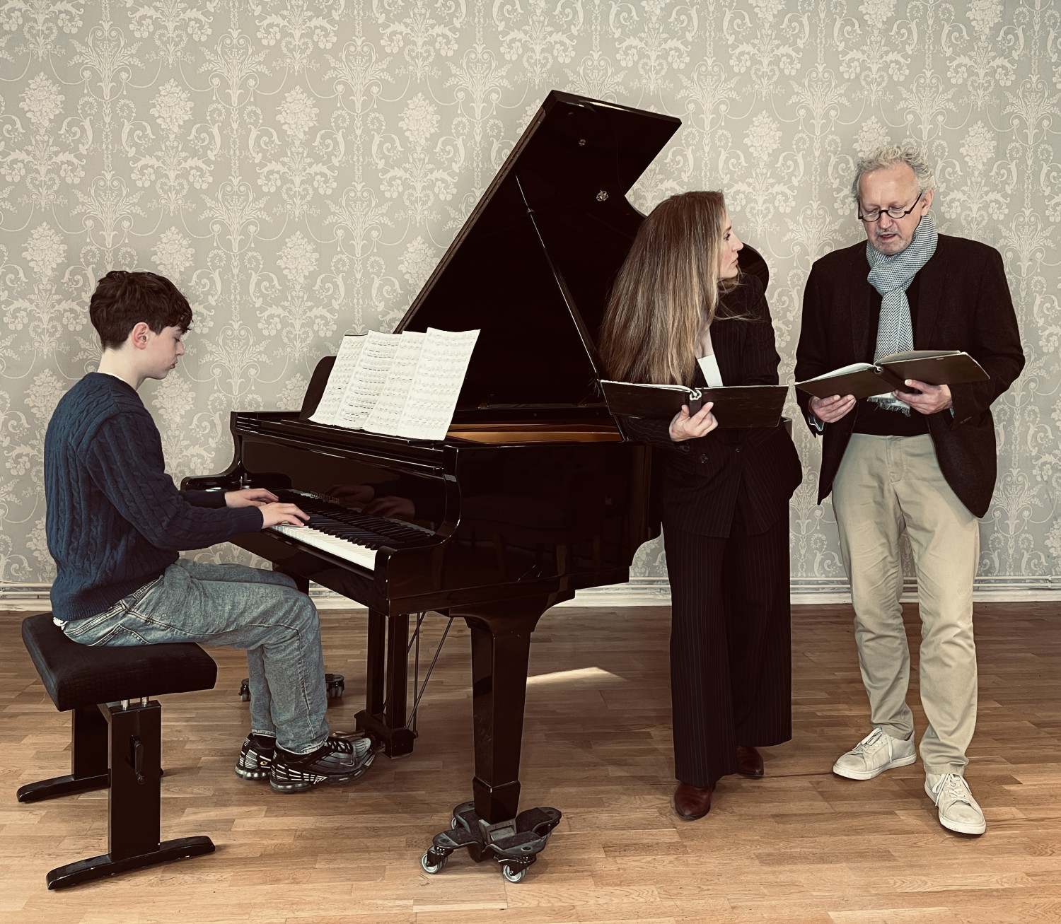 Eine Person am Klavier sitzend spielt, zwei Personen neben dem Klavier stehend lesen etwas vor