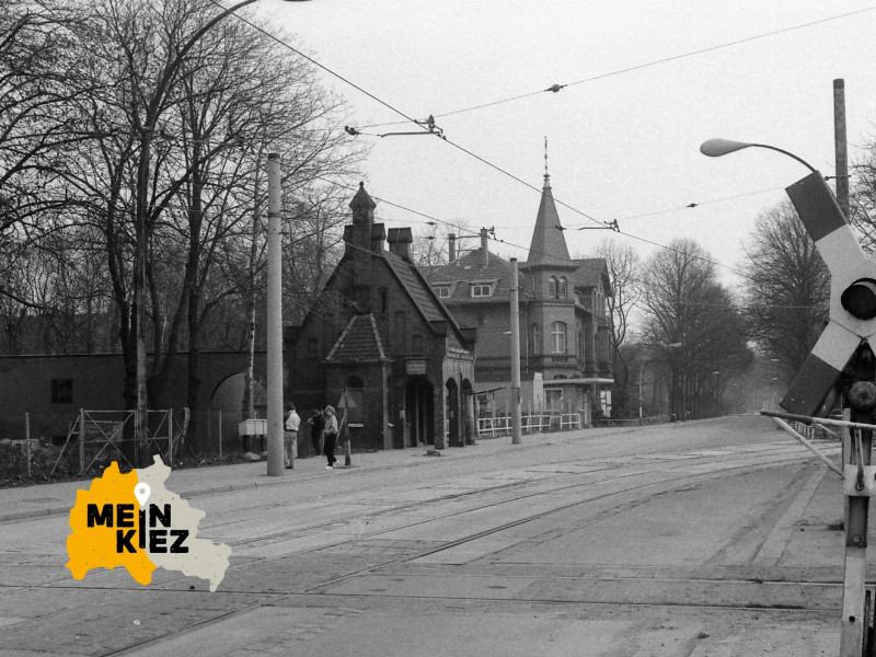 Schwarz-weiße Aufnahme einer Straßenbahnkreuzung mit Andreaskreuz und historischen Gebäuden im Hintergrund, zwei Personen gehen am Gehweg.