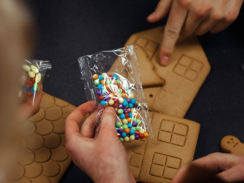 Kinderhände halten eine Packung mit bunten Perlen über die Einzelteile eines Lebkuchenhauses