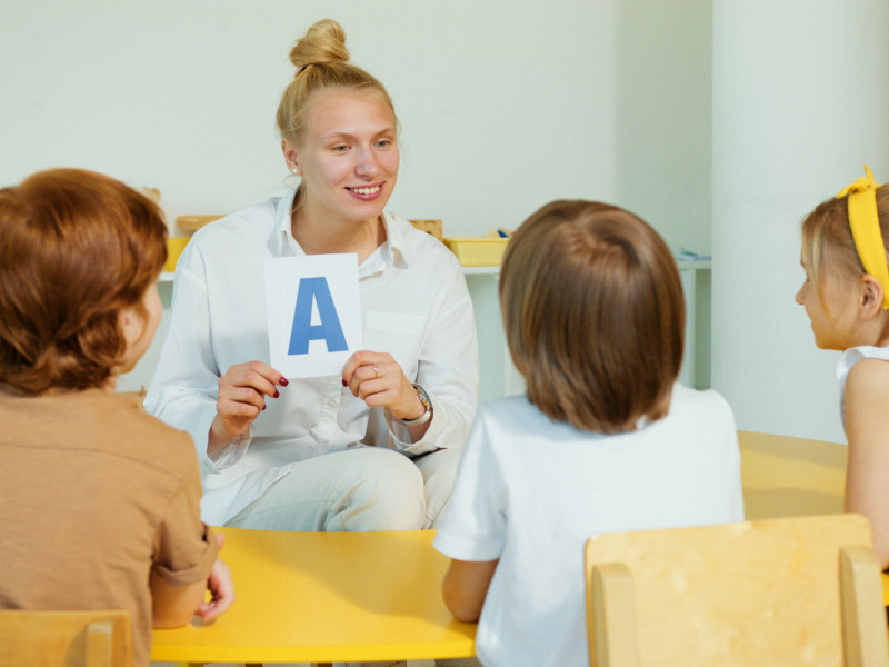 Eine erwachsene Frau zeigtv ein Blatt mit einem A hoch. Kinder sitzen um sie herum und schauen zu. 