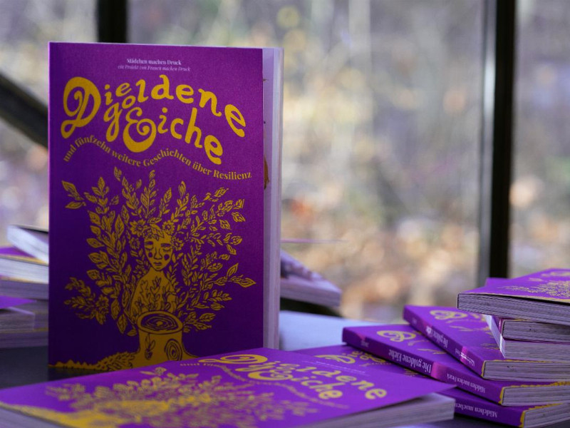 Mehrere lila Bücher mit goldener Schrift und Illustration mit dem Titel 'Die goldene Eiche' liegen auf einem Tisch vor einem Fenster mit unscharfem Waldhintergrund