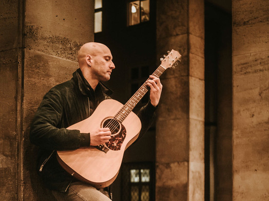 Ein Mann spielt Gitarre gelehnt an die Wand. Draußen.
