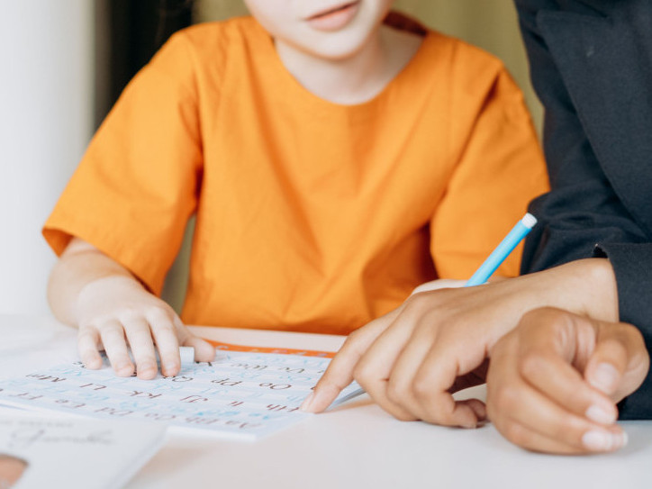 Ausschnitt ab den Schultern bis zum Unterkörper. Ein Kind und eine erwachsene Person sind geneigt über einen Tisch mit einem Blatt.rson