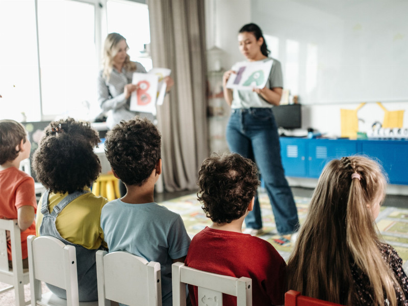 Zwei junge Frauen stehen vor fünf Kindern die auf Stühlen sitzen und den Damen zuhören.