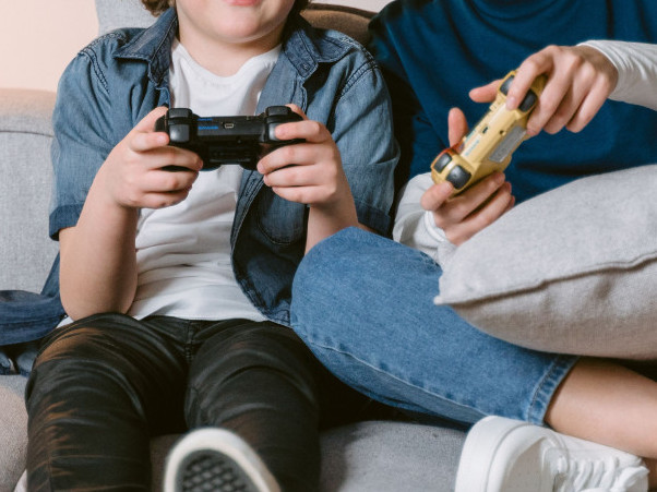 Ein Kind und eine erwachsene Person spielen mit zwei Controllern auf einem Sofa.