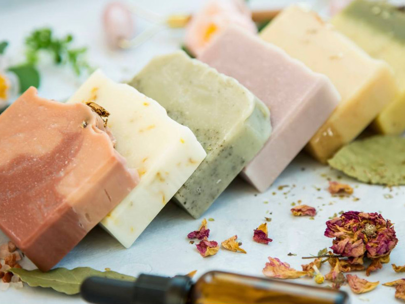 mehrere verschiedenfarbige geschnittene Seifen liegen gekippt auf einem Tisch, davor eine kleine getrocknete Rose mit verstreuten Blütenblättern
