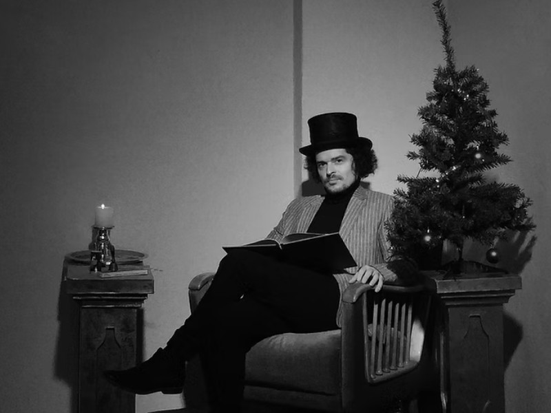 sw-Foto, ein Mann mit Zylinder sitzt auf einen alten Sessel mit einem Buch in der Hand, daneben steht ein Weihnachtsbaum