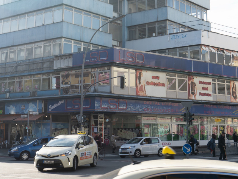 Straßenkreuzung an der Kurfürstenstraße mit Geschäftshaus, Autos und Passant*innen
