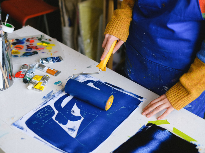 Eine Person druckt mit einem Farbroller blaue Farbe auf ein Papier. Ein Motiv entseht 