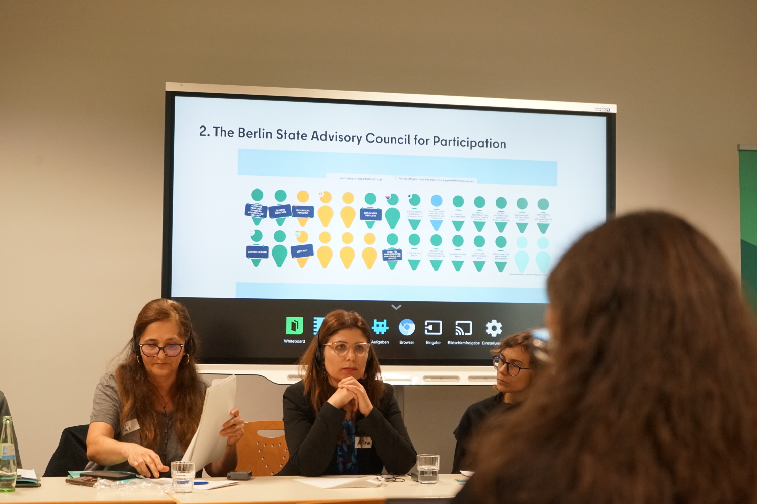Drei Frauen sitzen an einem Tisch und halten einen Vortrag. Im Hintergrund wird eine Präsentation auf einer Leinwand gezeigt.