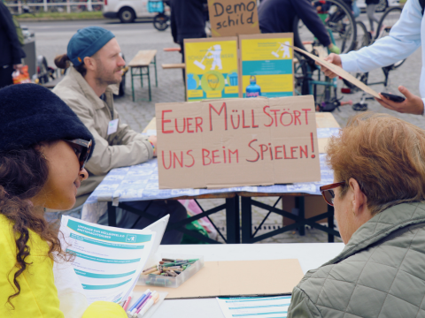 Menschen sitzen an Tischen beim Müllfest 2024. Auf einem Schild steht Euer Müll stört uns beim spielen.