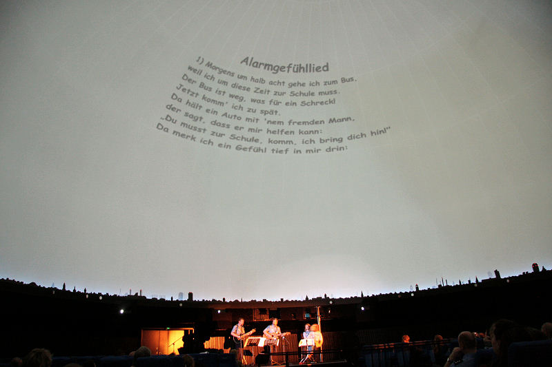 Bild von der Planetarium-Aufführung; Liedtext an der Decke und Bühne mit Darstellern