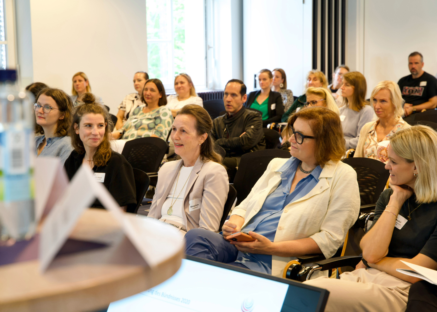 Ein Publikum aus mehreren Personen sitzt in Reihen in einem Konferenzraum und hört aufmerksam zu. Einige lächeln oder machen sich Notizen. Im Vordergrund ist unscharf ein Tisch mit Namensschild und Wasserflasche zu sehen.