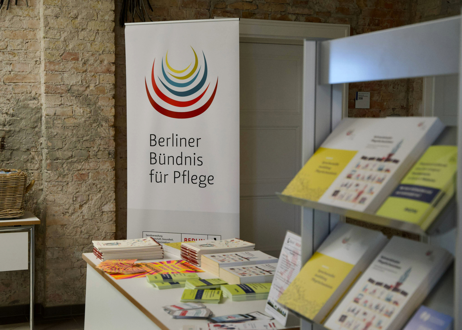 Ein Infotisch mit verschiedenen Broschüren steht vor einem Roll-up mit dem Logo und Schriftzug "Berliner Bündnis für Pflege". Rechts im Bild ist ein Broschürenständer mit weiteren Publikationen. Die Publikationen stehen im Flur, der eine Ziegelwand hat. Hinter dem Roll-up befindet sich eine weiße Tür.