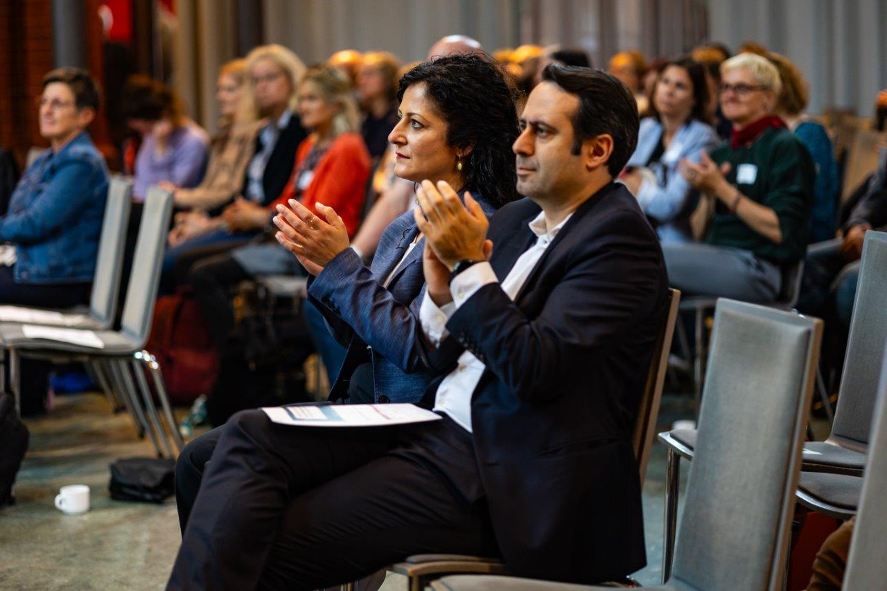 Sozialsenatorin Cansel Kiziltepe und Sozialstaatssekretär Aziz Bozkurt applaudieren im Publikum.