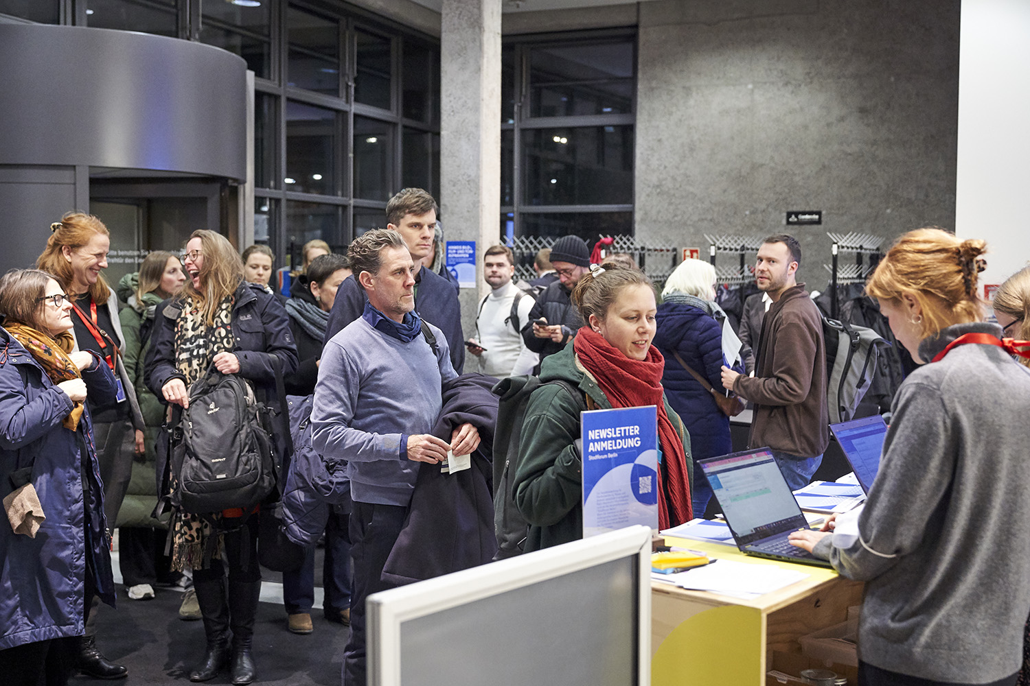 Personen stehen in einer Schlange vor einem Anmeldecounter. Eine Person tippt auf einem Laptop.