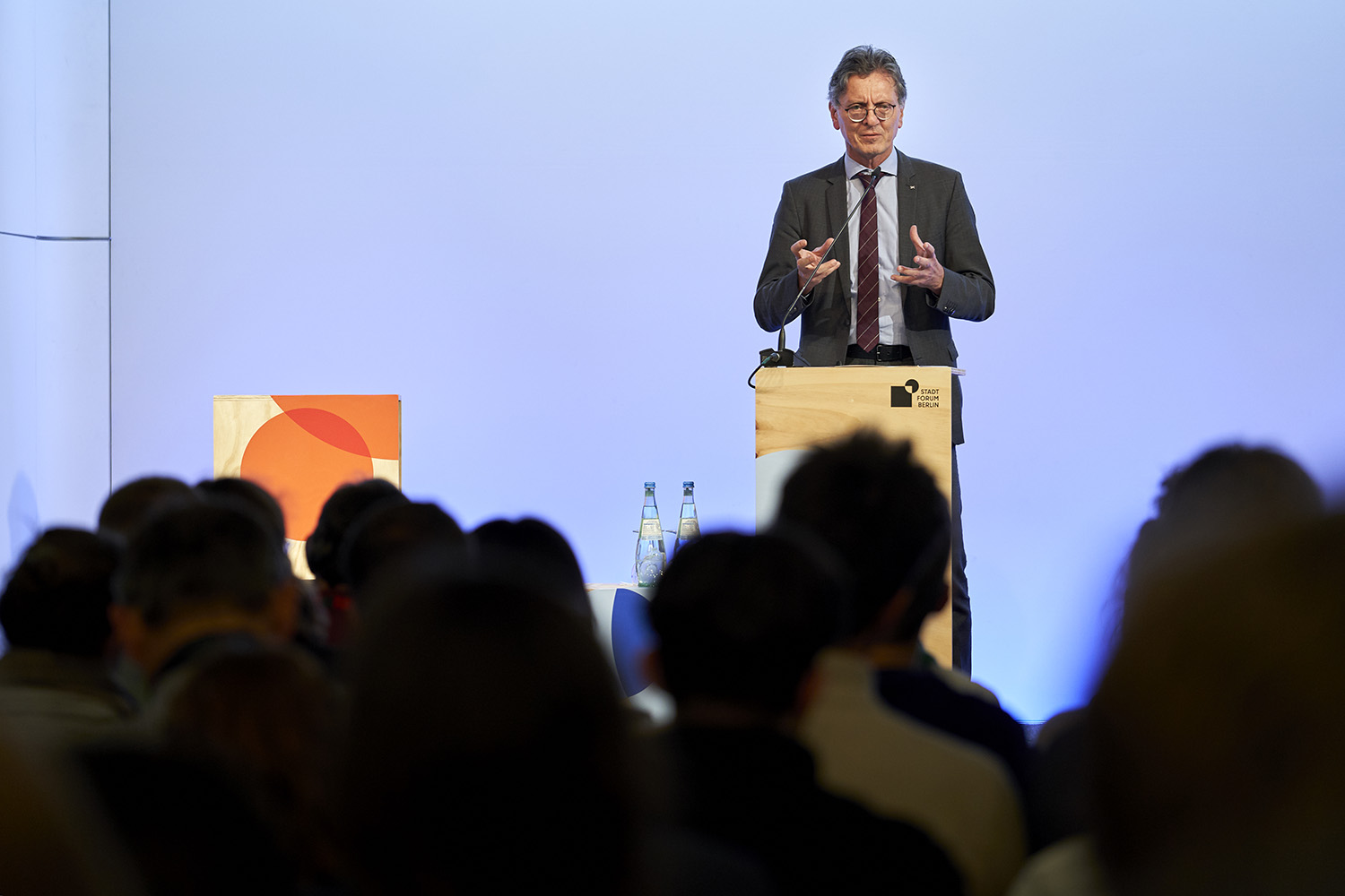 Ein Mann steht hinter einem Pult. Er spricht in ein Mikrofon und gestikuliert mit den Händen vor sich. Der Hintergrund ist Blau. Im Vordergrund sind die Köpfe des Publikums von hinten zu sehen.
