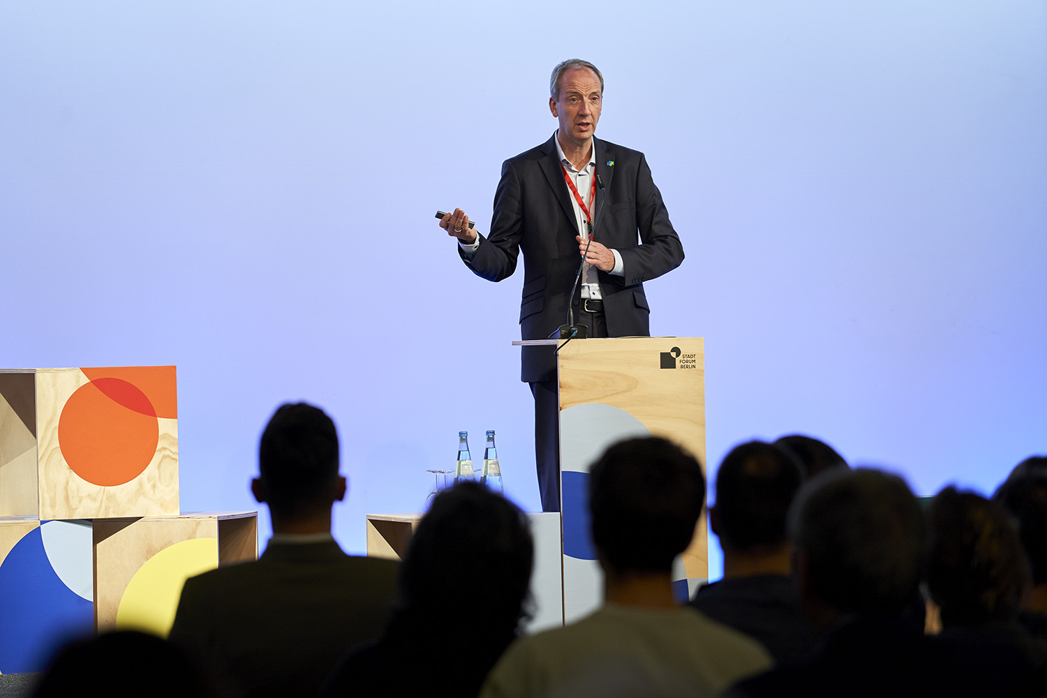 Ein Mann steht hinter einem Pult. Er spricht und zeigt mit einer Hand nach rechts. Auf der Bühne neben ihm stehen bunte Sitzmöbel. Vor ihm sieht man die Hinterköpfe des Publikums.