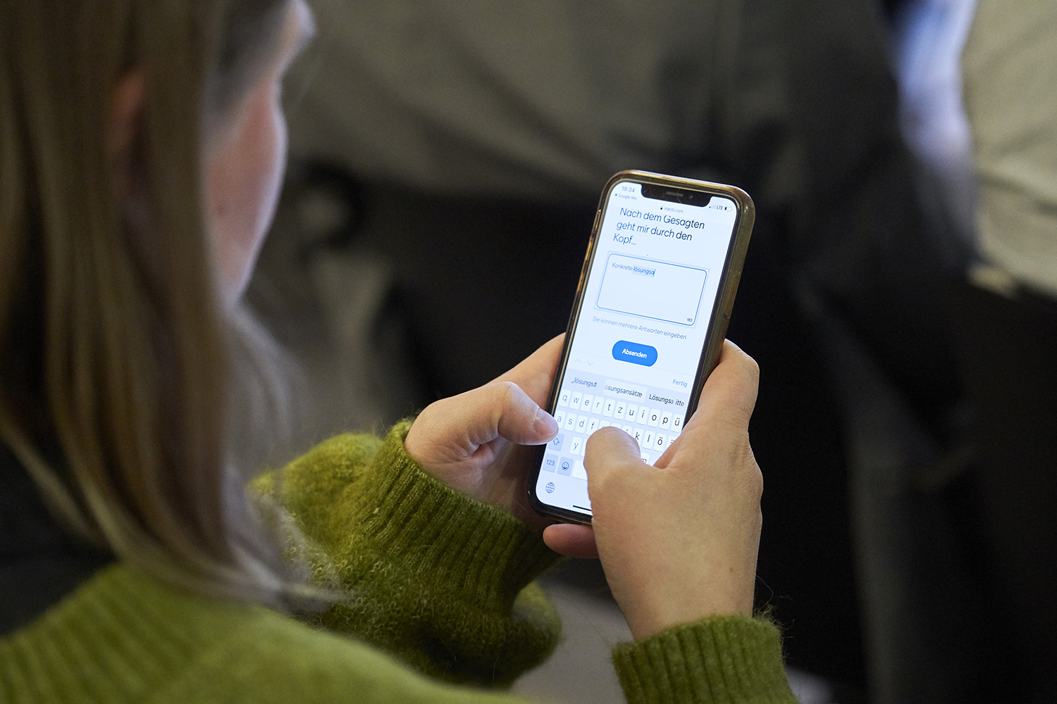 Eine Frau hält ein Handy in beiden Händen und tippt mit den Daumen. Auf dem Bildschirm ist die Oberfläche des Programms Mentimeter zu sehen.