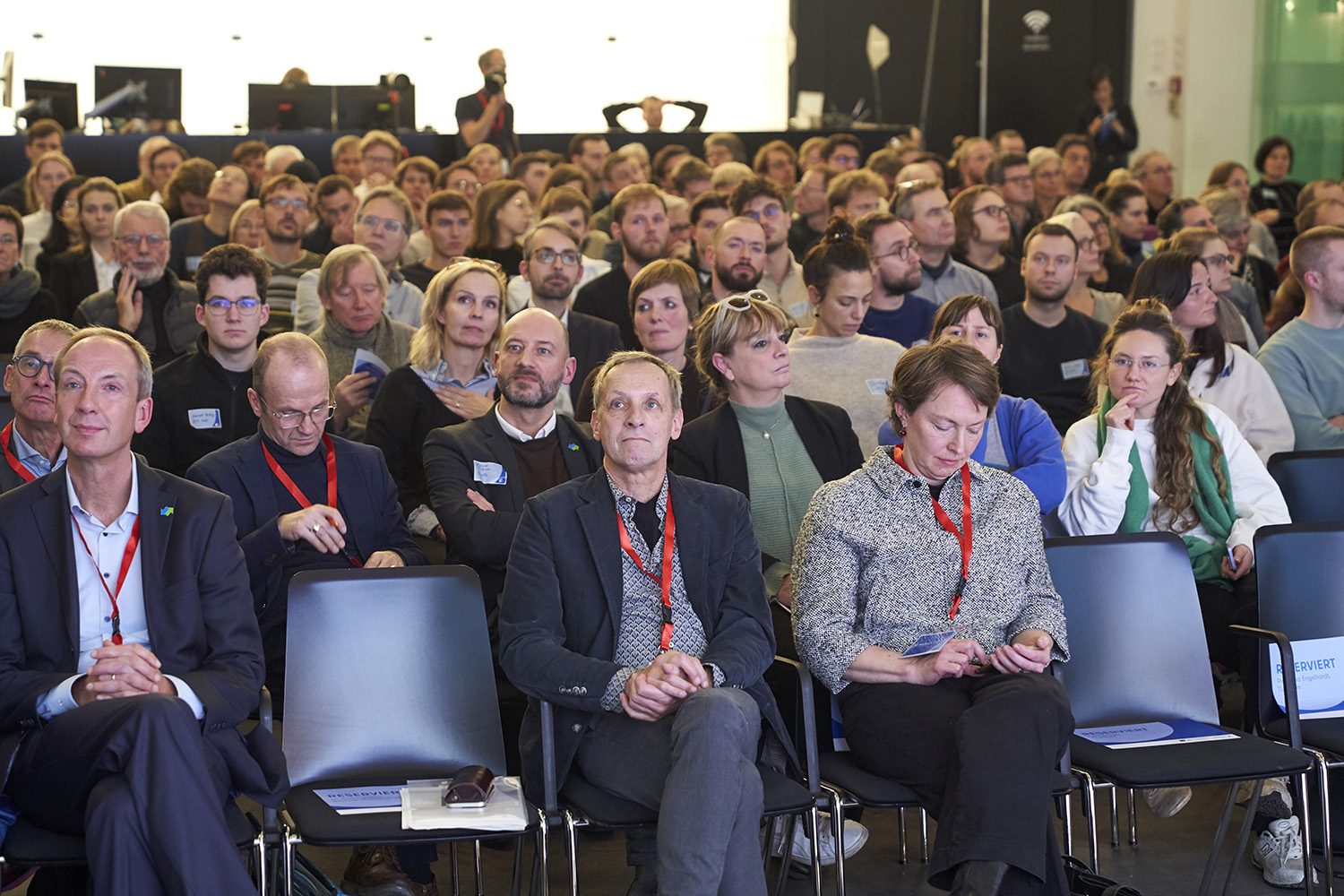 Viele Personen sitzen in Stuhlreihen und schauen der Kamera entgegen.