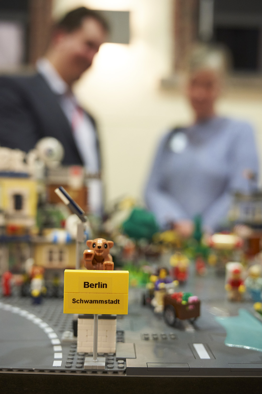 Vor einem Modell aus LEGO ist ein Ortsschild aus LEGO nachgebaut. Darauf steht „Berlin Schwammstadt“. Auf dem gelben Schild sitzt ein Bär. Im Hintergrund sieht man verschwommen zwei Personen.