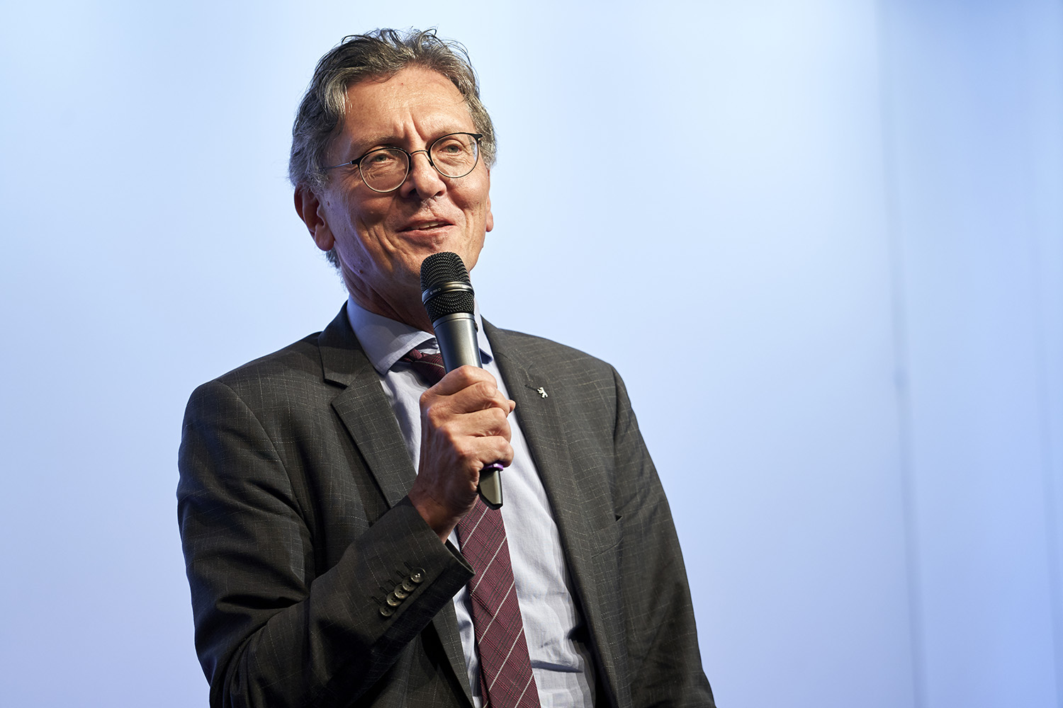 Ein Mann im Anzug und mit Brille spricht in ein Handmikrofon. Er steht vor einer blauen Bühnenleinwand.