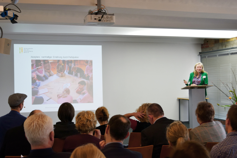Sabine Schulz-Greve, Vernetzungsstelle Kita- und Schulverpflegung Berlin hält einen Leinwand-Vortrag an einem Pult stehend vor Publikum