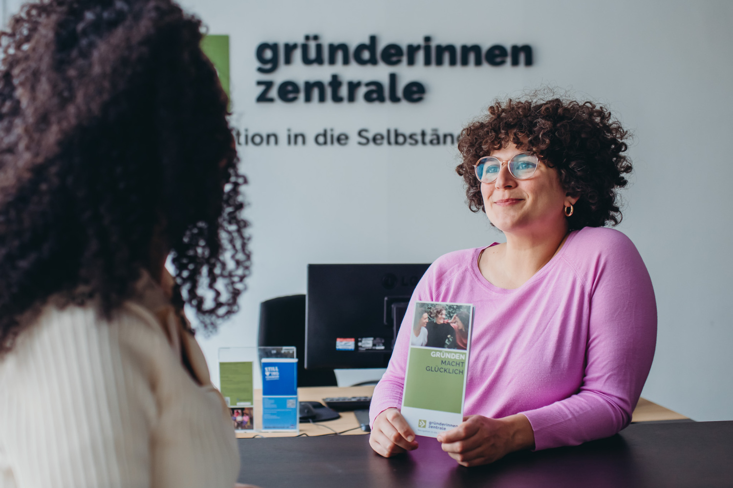 Eine Frau berät eine andere Frau und hält den Flyer der Gründerinnenzentrale hoch