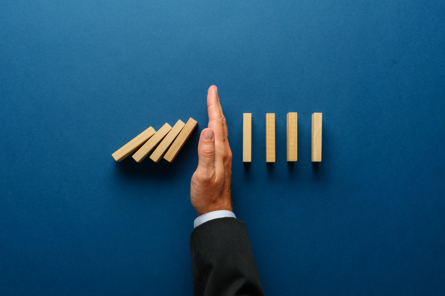 Hand in Anzugärmel stoppt fallende Dominosteine, daneben stehende Dominosteine auf blauem Untergrund.