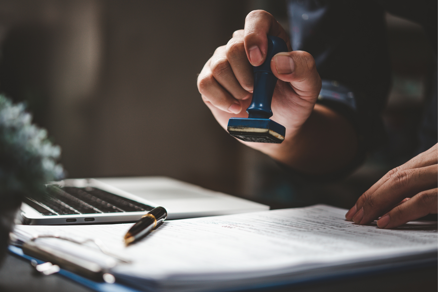 Hand hält einen blauen Stempel über einem Dokument auf einem Schreibtisch mit Kugelschreiber und Laptop.