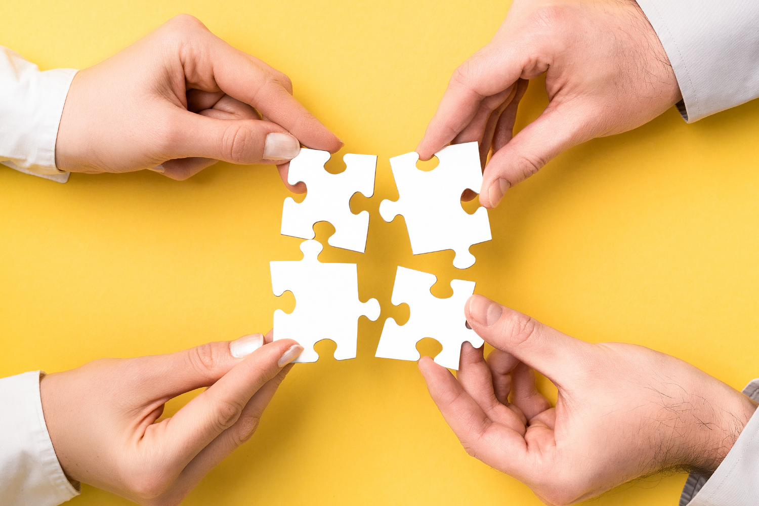 Vier Hände setzen vier weiße Puzzleteile auf gelbem Untergrund zusammen.