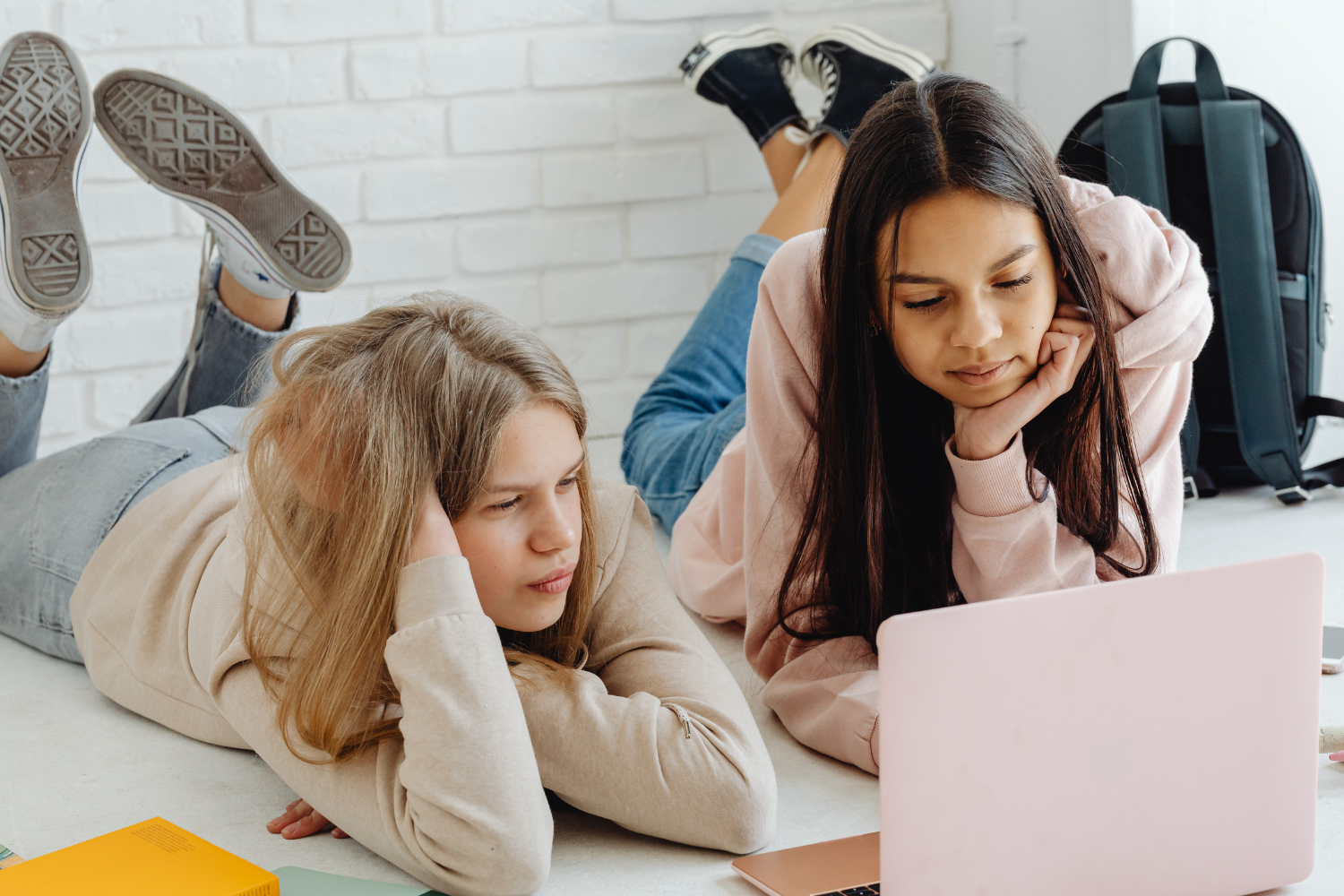 Zwei Jugendliche liegen auf dem Boden, schauen auf einen Laptop und haben einen Rucksack und ein Heft neben sich liegen.