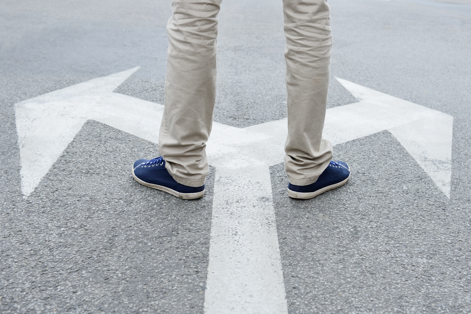 Beine einer Person auf einer Straße, vor zwei sich gegenüberstehenden Pfeilen, die eine Weggabelung symbolisieren.