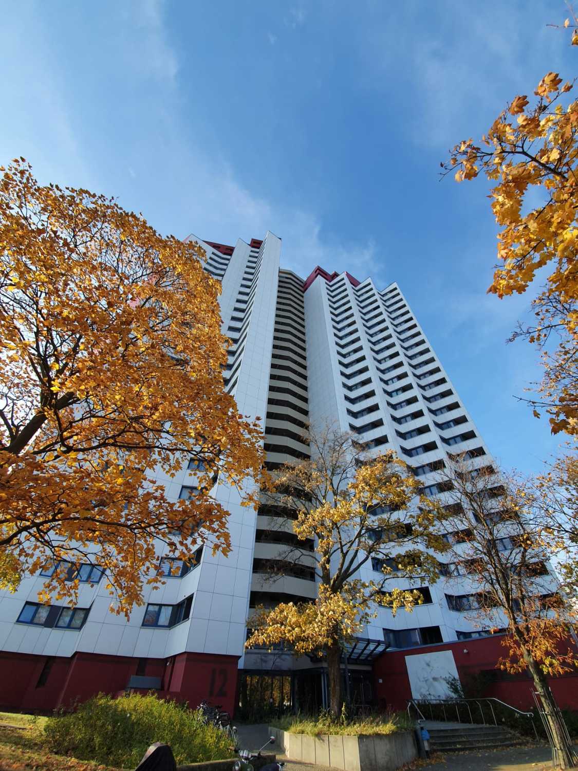 Ein Hochhaus. Der Sockel des Hauses ist dunkelrot. Die Fassade weiß. Man sieht die Fenster pro Etage. In der Mitte des Hauses ist das geschlossene Treppenhaus. Dieser Bereich ist grau gestrichen. Der Himmer ist blau mit leichten weißen Wolken. Rechts und links des Bildausschnittes stehen Laubbäume, deren Blätter orange gefärbt sind.
